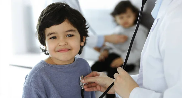 Médico-mujer examinando a un niño paciente por estetoscopio. Lindo chico árabe y su hermano en la cita con el médico. Concepto de medicina —  Fotos de Stock