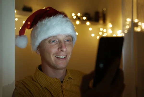 Gülümseyen genç adam Noel Baba şapkası takıyor, bakıyor ve arka planda Noel ışıkları olan akıllı bir telefon kullanıyor..
