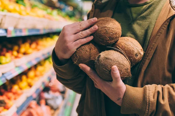 A male shopper in a supermarket holds coconuts in his hands