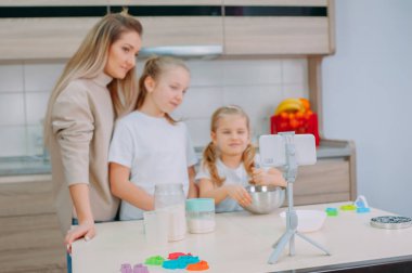 Annem kızlarına mutfakta hamur pişirmeyi öğretiyor. Aile akıllı telefondan aşçılık videosu çekiyor.