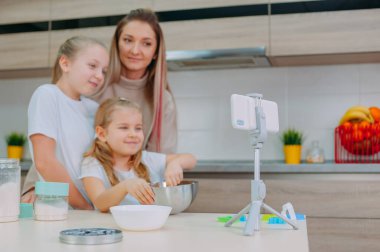 Annem kızlarına mutfakta hamur pişirmeyi öğretiyor. Aile akıllı telefondan aşçılık videosu çekiyor.