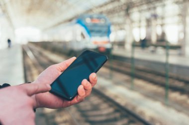 Bir adam, tren istasyonundaki bir trenin arka planında akıllı telefonun maketini tutuyor.