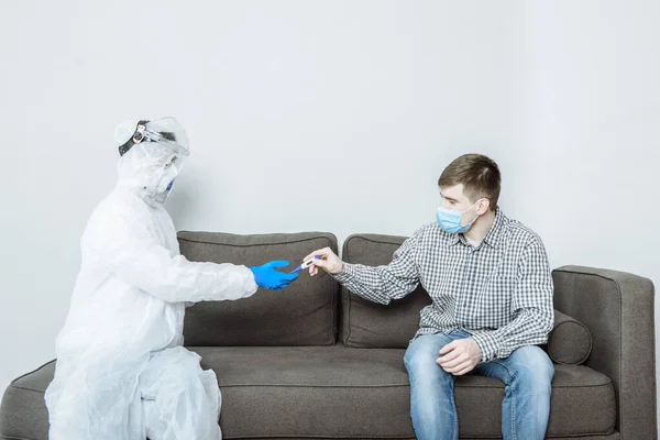 a doctor in a protective suit PPE Hazmat wearing a mask and glasses gives a thermometer to a patient for a health check in case of symptoms of the coronavirus covid-19