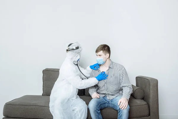 A doctor in a protective suit PPE hazmat in a mask and gloves examines the patient and listens with a stethoscope to breathing with symptoms of coronavirus covid-19