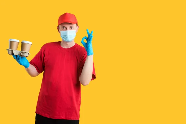 Home delivery of goods. Delivery man in a red cap and T-shirt wearing a mask and gloves with eco cups of coffee shows an okay sign on a yellow background isolate. copy spaces