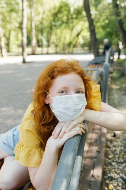 Tek kullanımlık maskeli kızıl saçlı kız şehir parkında yürüyor..