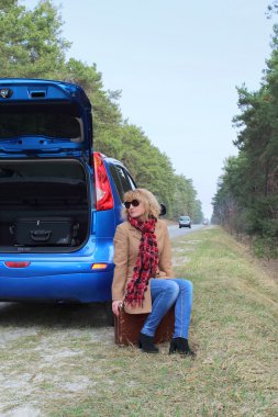 Mavi araba yakınındaki yolda bavul ile oturma sevimli Bayan