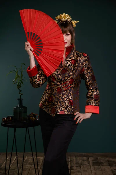 Krásná žena s bystrým make-up pózování v čínském kostýmu v minimalistickém interiéru s ventilátorem a dýkou — Stock fotografie