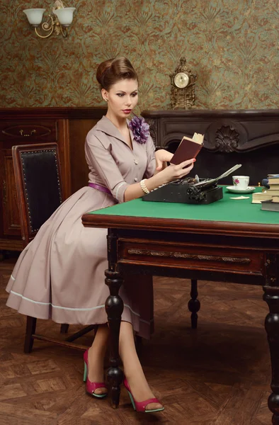 Pin Up beautiful young woman in vintage interior reading a book and prints on an old typewriter — Stock Photo, Image