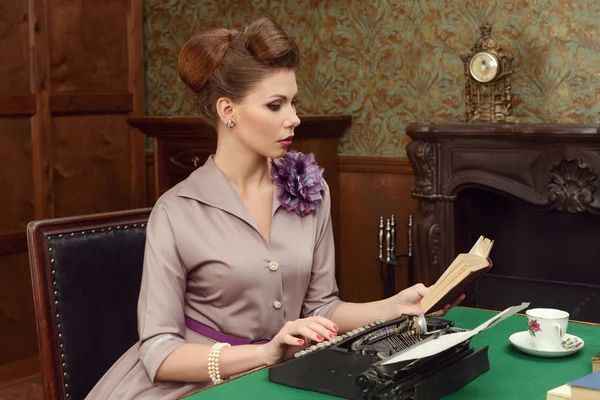 Pin Up beautiful young woman in vintage interior reading a book and prints on an old typewriter — Stock Photo, Image