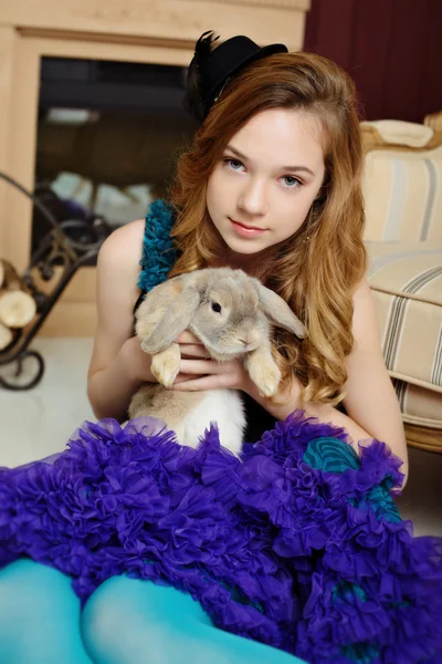 A young girl in the image of Alice in Wonderland is sitting near the fireplace and holds the rabbit