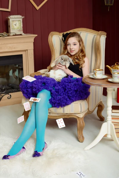 Young girl in the image of Alice in Wonderland is sitting in a chair near the fireplace and holds a rabbit — Stock Photo, Image