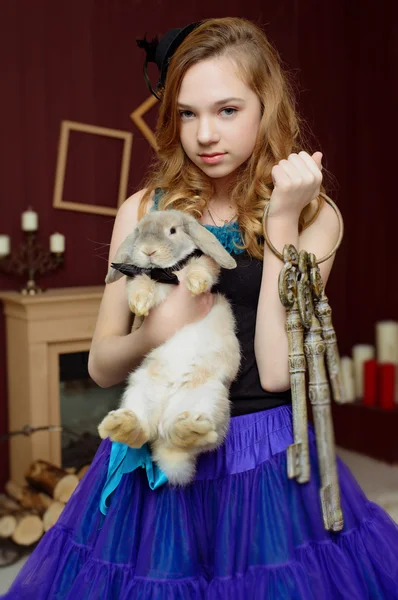 Young girl in the image of Alice in Wonderland stands near the fireplace and holds a rabbit and a bunch of keys