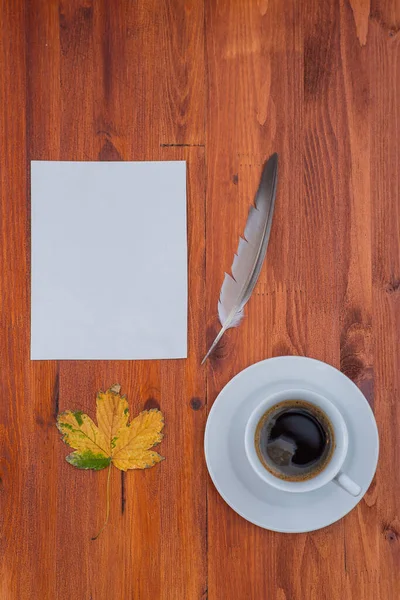 wood table. plain white sheet of paper, cup of coffee. colorful dry leaf. bird feather. autumn background.