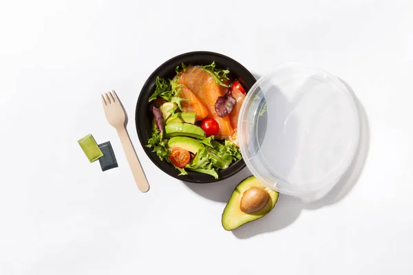 take away box with salmon salad an avocado and fork on white background