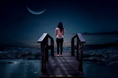 Genç bir kadın sabahın erken saatlerinde ahşap bir köprüde tek başına duruyor. Yarım ay gecesi.