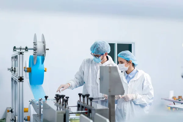 Staff and workers at the mask factory are monitoring the operation of the machines. A mask is being produced on machine. Factory workers are discussing.