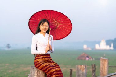 Genç bir Burmalı kadın Asya 'nın en uzun meşe köprüsünde kırmızı bir şemsiyeyle yürüyor. U-bein Köprüsü, Mandalay, Myanmar. Metin için boşluk.