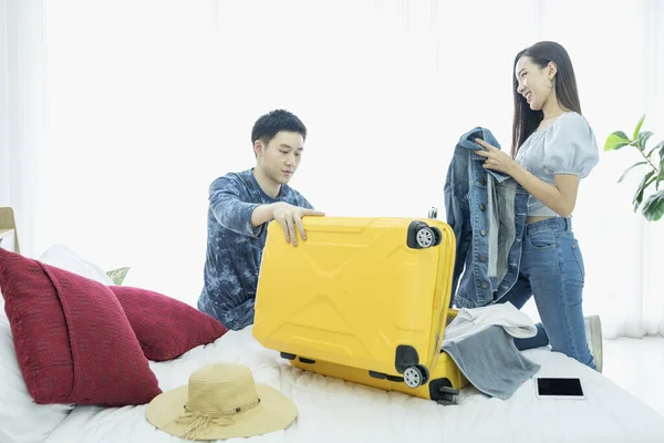 Young Couple are preparing to travel. Happy young friends packing clothes put in suitcase on bedroom. Female and Male using computer Tablet planning travel information.