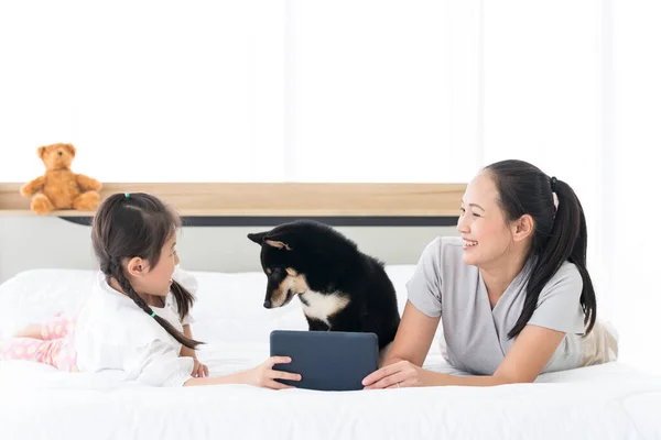 幼い母親と小さな娘がマルタ犬とベッドで遊んでいます 幸せな母親保持彼女の子供使用タブレットとともに犬でベッドルーム — ストック写真