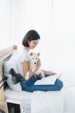 Beyaz bir Shiba Inu ile yatakta kitap okuyan genç bir kadın. Kız ve beyaz Hokkaido inu köpeği.