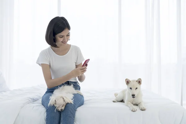 若いアジアの女性は携帯電話で写真を撮る 若いアジアの女性と少女は犬とベッドにいた 女の子と白 北海道犬とマルタ犬 — ストック写真