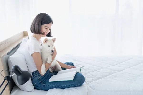 寝室に白い芝犬とベッドの上で本を読んでいる若い女性 女の子と白 北海道犬 — ストック写真