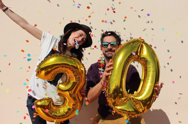 Coppia allegra celebra un compleanno trentennale con grandi palloncini d'oro e piccoli pezzi di carta colorati in aria . — Foto Stock