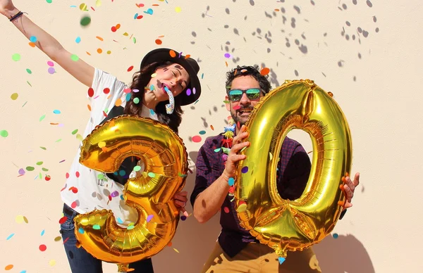 Coppia allegra celebra un compleanno trentennale con grandi palloncini d'oro e piccoli pezzi di carta colorati in aria . — Foto Stock