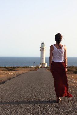 Young beautiful woman walking slowly to Cap de Barberia's lighthouse during an amazing summer sunset. clipart
