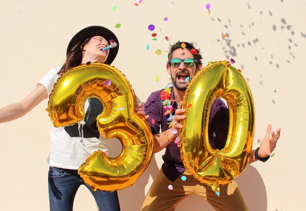 Χαρούμενο ζευγάρι γιορτάζει τριάντα χρόνια γενέθλια με μεγάλη χρυσή μπαλόνια και πολύχρωμα μικρά κομμάτια χαρτιού στον αέρα. — Φωτογραφία Αρχείου