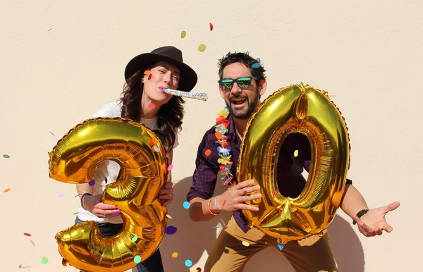 Veselý pár slaví třicet let narozeniny s velkými zlatými balónky a barevné kousky papíru ve vzduchu. — Stock fotografie
