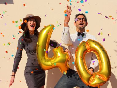 Cheerful couple celebrates a forty years birthday with big golden balloons and colorful little pieces of paper in the air. clipart