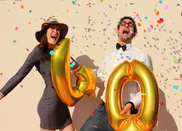 Alegre pareja celebra un cumpleaños de cuarenta años con grandes globos dorados y pequeños trozos de papel coloridos en el aire . — Foto de Stock