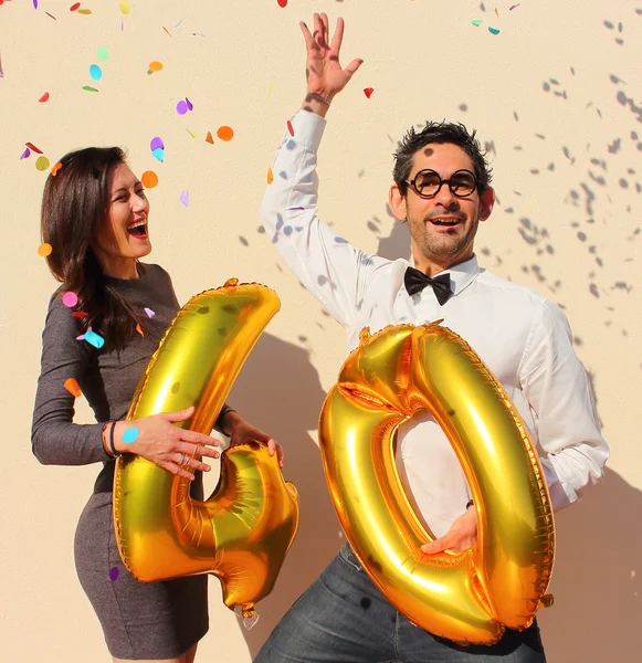 Coppia allegra celebra un compleanno di quarant'anni con grandi palloncini d'oro e piccoli pezzi di carta colorati in aria . — Foto Stock