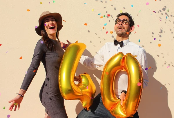 Alegre pareja celebra un cumpleaños de cuarenta años con grandes globos dorados y pequeños trozos de papel coloridos en el aire . — Foto de Stock