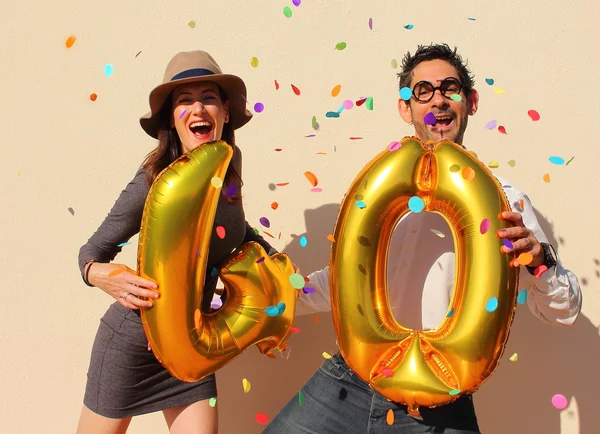 Alegre pareja celebra un cumpleaños de cuarenta años con grandes globos dorados y pequeños trozos de papel coloridos en el aire . — Foto de Stock