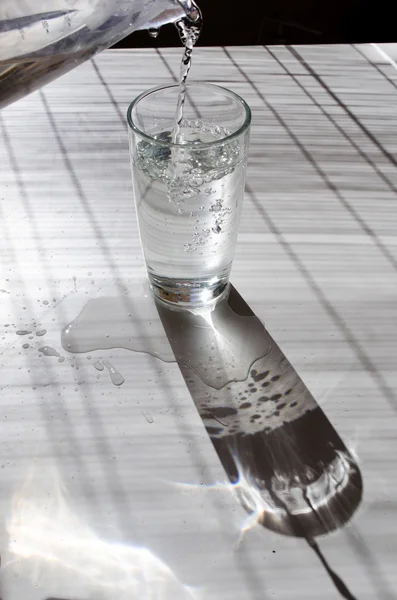 Verter agua en un vaso aligerado con sol sobre una mesa blanca — Foto de Stock