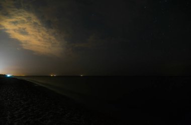 Güzel yıldızlı gece manzarası. Gökyüzünde parlayan yıldızlar ve Azov Denizi kıyısındaki kumlu plajlar. Dalgalar kıyıya vuruyor ve ufukta Mariupol şehrinin uzak ışıkları görünüyor. Villalar, kulübeler ve sahildeki evler.