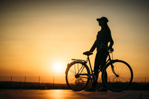 Beautiful hipster girl with a bicycle on the road during sunset — Stock Photo, Image