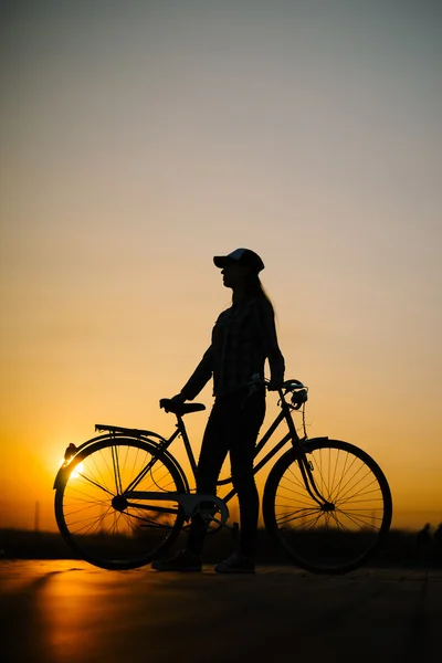 Linda garota hipster com uma bicicleta na estrada durante o pôr do sol — Fotografia de Stock