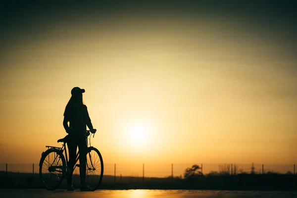 Linda garota hipster com uma bicicleta na estrada durante o pôr do sol — Fotografia de Stock