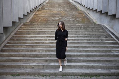 Fashion portrait of a sexy brunette woman with a make up wearing black clothes. She is wearing black modern dress, white sneakers and glasses