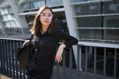 Business woman portrait in city streets at sunset. Sexy brunette wearing black dress, glasses and black leather bag outdoor
