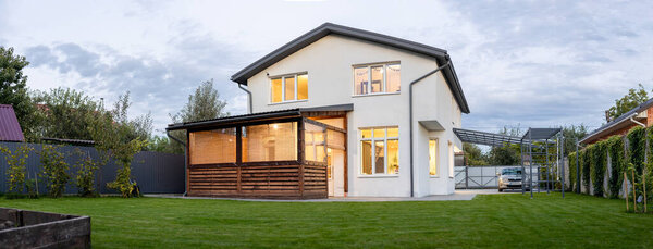 Exterior view of modern white house with courtyard, patio area, green grass lawn, garden and a car in the evening