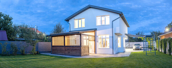 Exterior view of modern white house with courtyard, patio area, green grass lawn, garden and a car in the evening