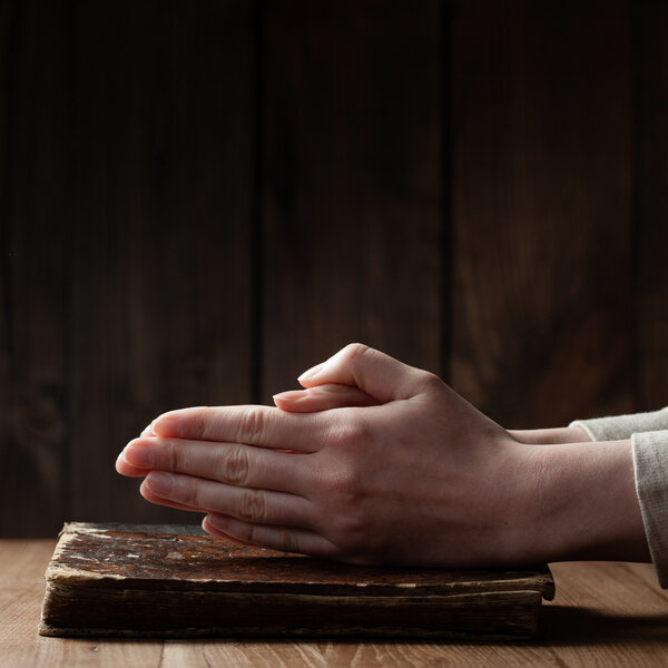 Руки женщины молятся с библией в темноте над деревянным столом
