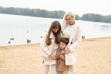 Kadın çocuk kumsalda soğuk havada yürür, annesi oğlu ve kızı hafif giysiler içinde. Birlikte iyi eğlenceler. Şefkat kavramı, sevgi, güzel aile ilişkilerini kucaklamak. Açık hava fotoğrafı.