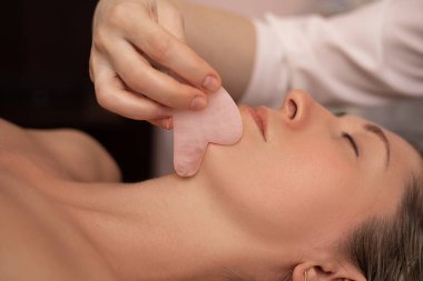 Geleneksel Çin masajı sırasında genç ve güzel bir kadın, Gua Sha. Yakın plan fotoğraf. SPA salonunda güzellik tedavisi. Yaşlanma önleyici cilt bakımı