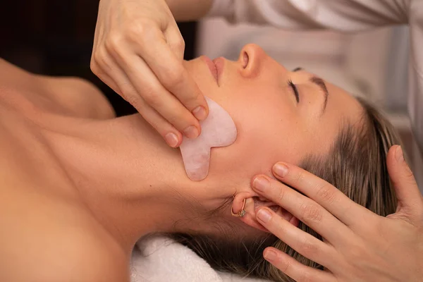 Young and beautiful woman during Chinese traditional massage - Gua Sha with stone. Close-up photo. Beauty treatment in SPA salon. Anti-aging skin care